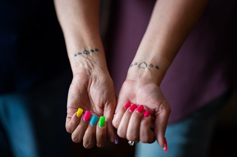 Two hands extended forward, showing wrists with matching heart tattoos in a row, with the central heart being outlined and flanked by smaller, filled hearts. The nails are painted in vibrant, multicolored shades, with one hand featuring yellow, pink, green, blue, and purple nails, and the other sporting bright pink nails.