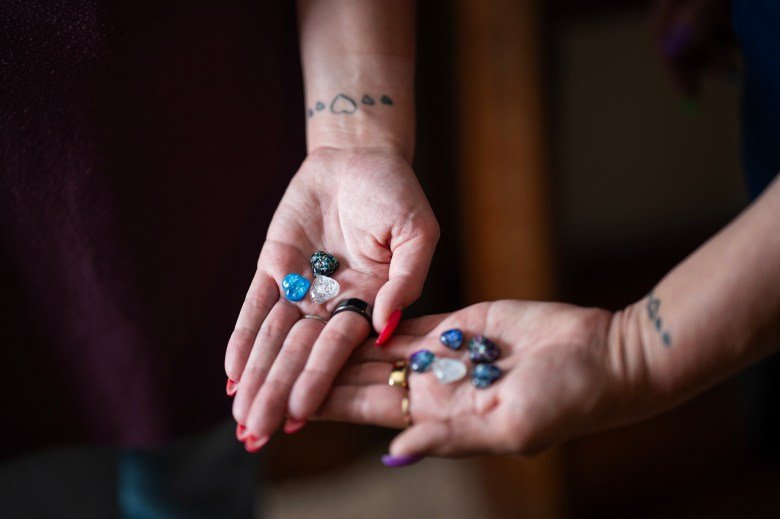 Two hands extended, each holding small, glittery, colored stones. Both wrists have matching tattoos of small, filled-in heart shapes. The stones are various shades of blue, white, and black, with a shiny, textured surface.