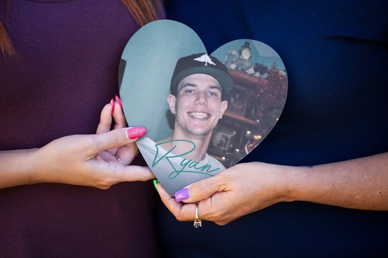 Two hands with painted nails hold a heart-shaped photograph of a person smiling and wearing a baseball hat. The hands belong to a mother and a daughter, and the image has a feeling of "remembrance." The image is focused on the heart and the hands.