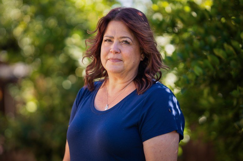 A person in a blue shirt stands in outdoors and in front of greenery. They look intently into the camera. A silver necklace rests on their chest.