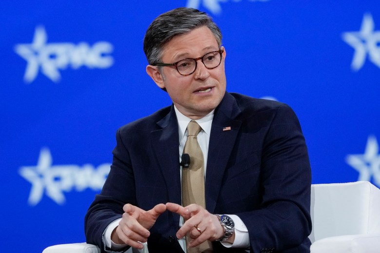 A person in a dark suit, white shirt, and beige tie speaks while seated on a stage with a blue backdrop featuring the CPAC logo. They wear glasses and have short, neatly combed hair. A small American flag pin is visible on their lapel, and a microphone is clipped to their tie. Their hands are gesturing as they speak.