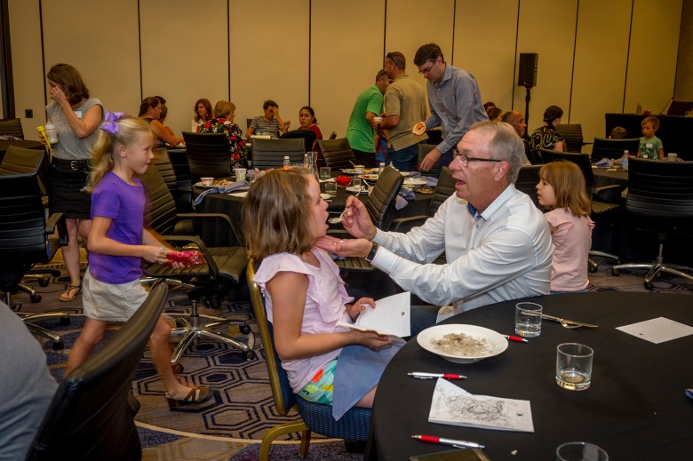 Amidst a busy hotel hall room filled with people and rounded black tables, a dad uses a spoon to feed his daughter.