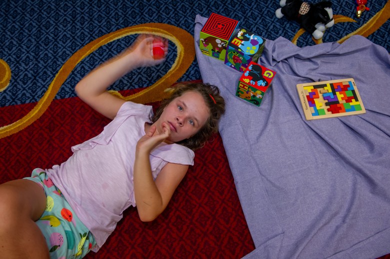 A young girl lays on a red and blue carpeted floor next to a purple sheet with toys and puzzels. She looks at the camera above her while holding a small red tennis ball.