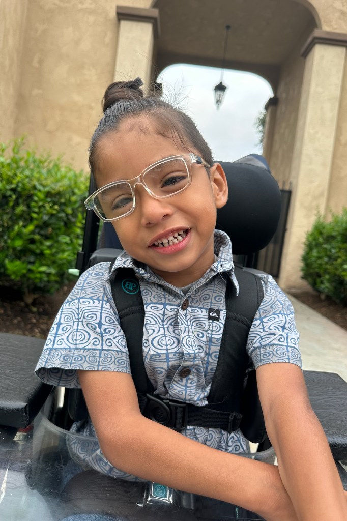 A child, sitting on a wheel chair and wearing clear glasses with a blue and white patterned shirt, tilts their head to their right as they smile at the camera.