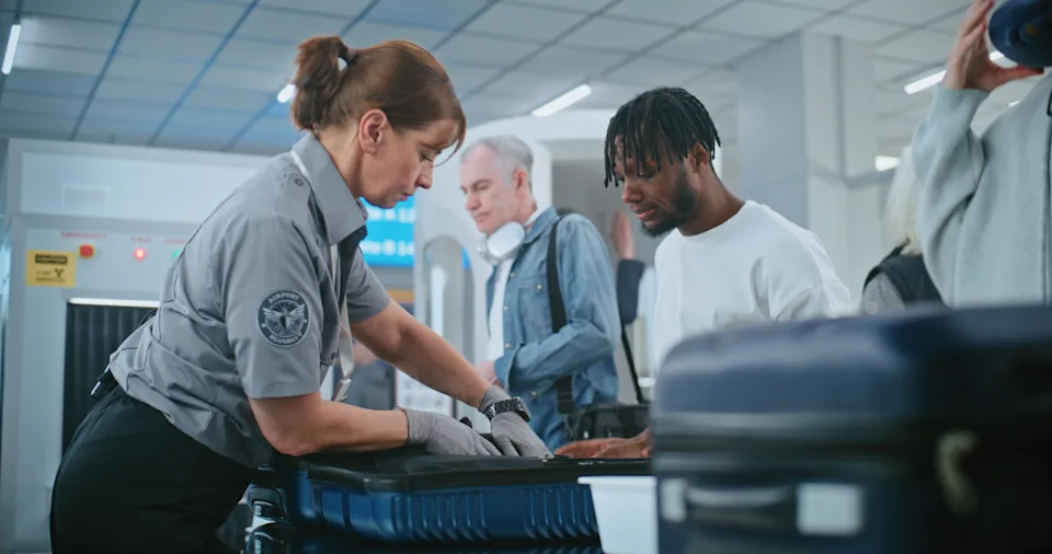 Security Checkpoint in Airport Terminal: Female TSA Worker Inspecting Baggage of Passenger before Boarding Flight, Finding and Confiscates Liquid. Queue of Diverse People During Screening Procedures.