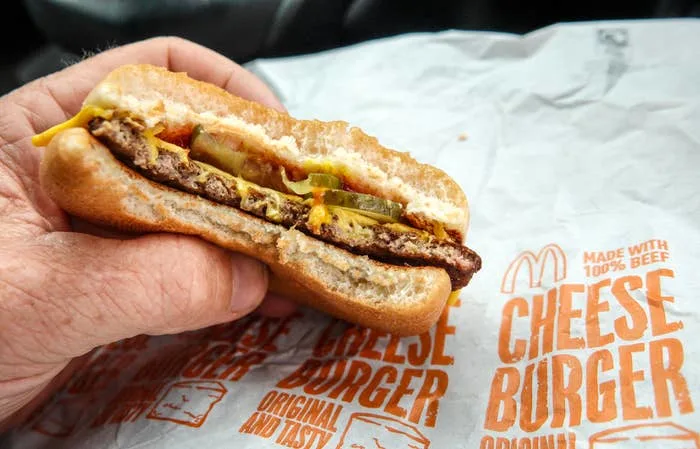 A person holding a McDonald's cheeseburger with visible layers of meat, cheese, pickles, and sauce, over branded packaging with text
