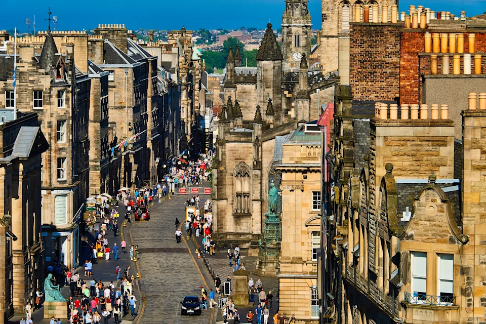 A bustling street in a historic city, lined with old stone buildings. Crowds of tourists explore the area on a sunny day