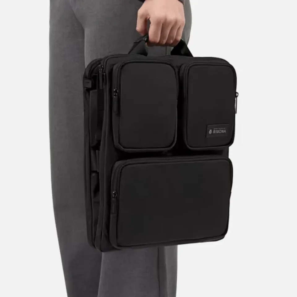 Black multi-pocket briefcase with top handle held by a person wearing gray pants.