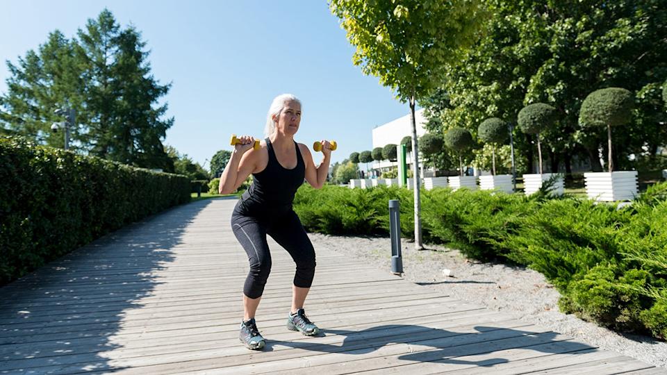 Senior woman performing a squat exercise with light dumbbells during an outdoor workout on a sunny day.
