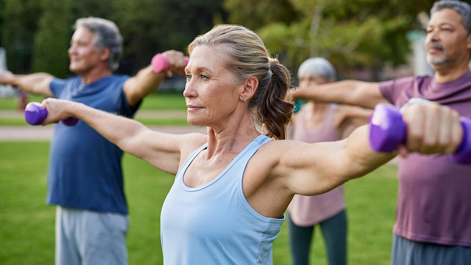 Active seniors performing shoulder raises with dumbbells during an outdoor group fitness class in a park.